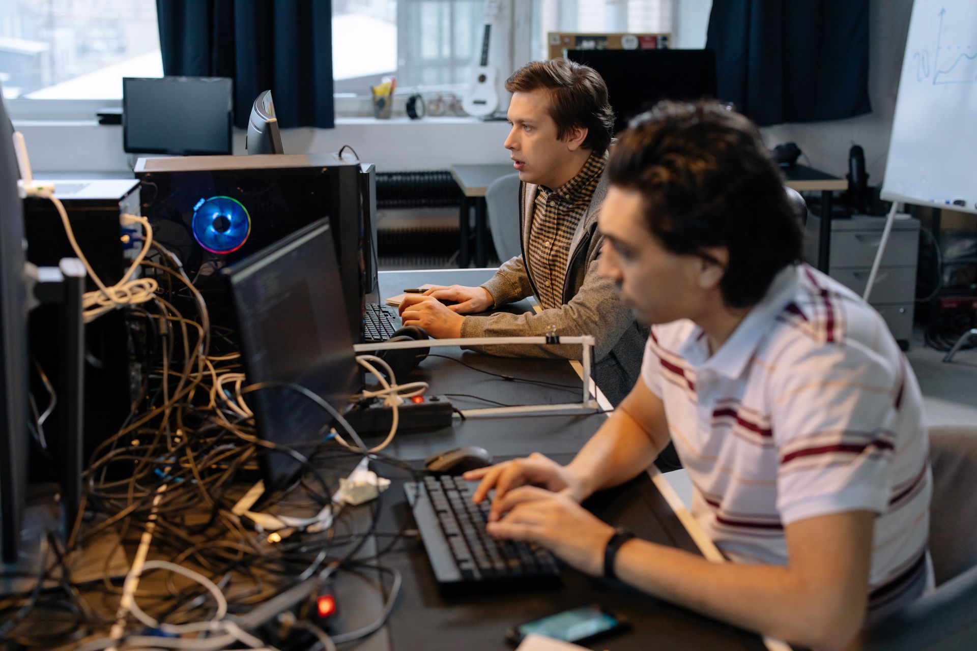 Zwei junge Männer sitzen nebeneinander an einem vollgestellten Schreibtisch und arbeiten konzentriert an Computern. Auf dem Tisch liegen zahlreiche Kabel, Tastaturen, Mäuse und Monitore. Ein PC-Gehäuse mit blau leuchtendem RGB-Lüfter ist sichtbar. Im Hintergrund ist ein Whiteboard sowie eine Gitarre an der Wand zu erkennen. Die Atmosphäre wirkt wie ein technisches Arbeits- oder Hackspace-Umfeld.