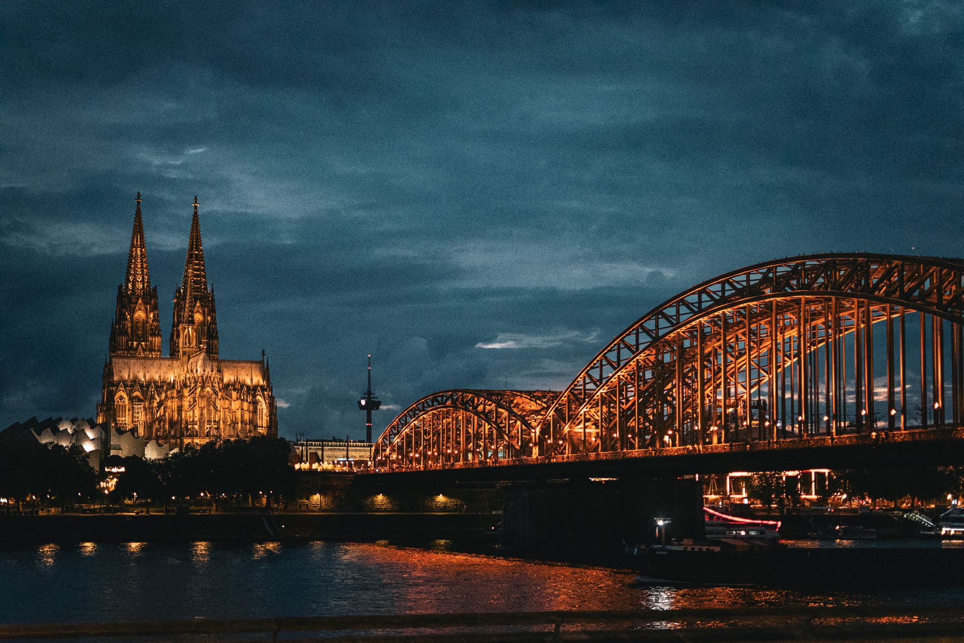 Nächtliche Stadtansicht von Köln mit dem beleuchteten Kölner Dom und der Hohenzollernbrücke über den Rhein. Der gotische Dom mit seinen zwei markanten Türmen erstrahlt in warmem Licht vor einem dramatisch bewölkten, blaugrauen Abendhimmel. Die Stahlbogenbrücke ist ebenfalls golden beleuchtet und spiegelt sich im Rhein. Im Hintergrund ist der Colonius-Fernsehturm zu erkennen. Rechts fährt ein beleuchtetes Schiff auf dem Fluss.