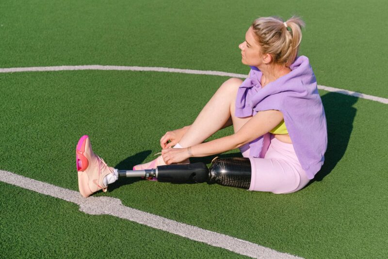 Eine junge Frau mit blonden Haaren sitzt auf einem grünen Kunstrasenplatz und bindet ihren Sportschuh. Sie trägt ein lilafarbenes Oberteil, pinke Shorts und ein gelbes Sport-Top. Ihr linkes Bein ist eine Unterschenkelprothese aus Karbonfaser. Die Szene wirkt entspannt und sportlich, aufgenommen bei Sonnenlicht.