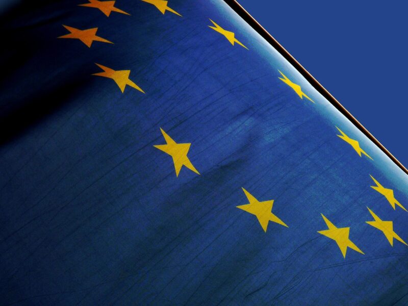 Nahaufnahme der Flagge der Europäischen Union. Der blaue Stoff mit dem Kreis aus zwölf goldenen Sternen füllt das gesamte Bild aus und ist leicht in Bewegung. Durch das Gegenlicht und die Falten des Stoffs wirken die Farben intensiv und kontrastreich. Im Hintergrund ist ein klarer blauer Himmel zu sehen.