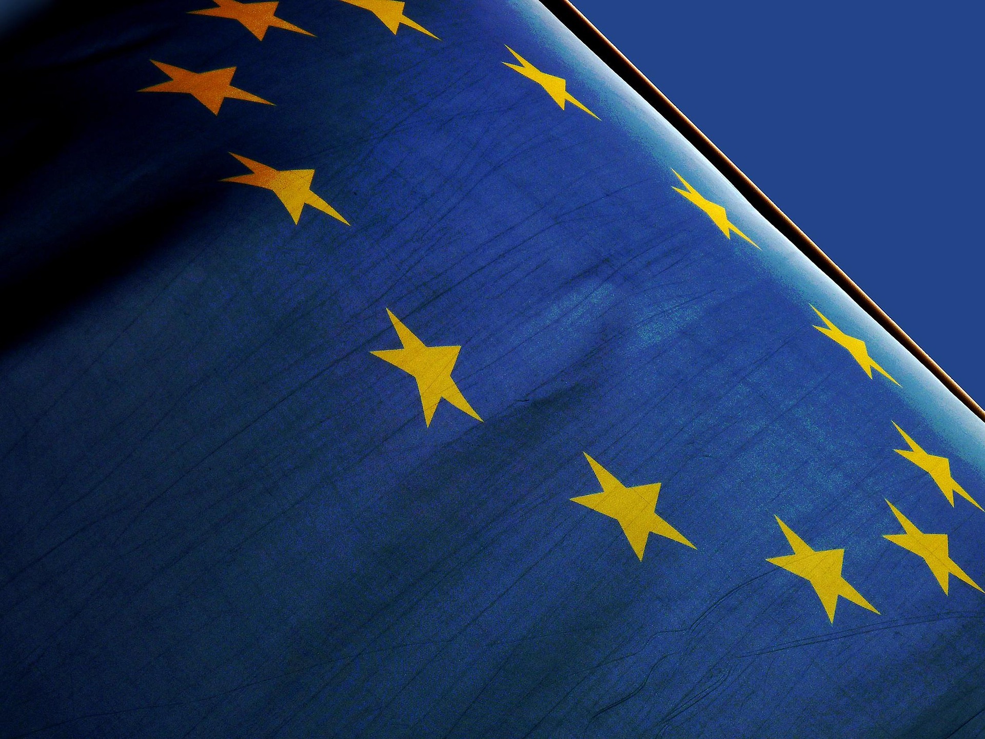 Nahaufnahme der Flagge der Europäischen Union. Der blaue Stoff mit dem Kreis aus zwölf goldenen Sternen füllt das gesamte Bild aus und ist leicht in Bewegung. Durch das Gegenlicht und die Falten des Stoffs wirken die Farben intensiv und kontrastreich. Im Hintergrund ist ein klarer blauer Himmel zu sehen.