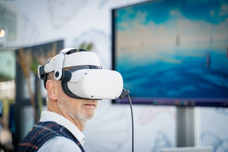 Ein älterer Mann mit grauem Bart trägt eine weiße VR-Brille und schaut in eine virtuelle Umgebung. Er ist von der Seite zu sehen und trägt ein weißes Hemd und eine karierte Weste. Im Hintergrund ist ein Bildschirm mit einer blauen, landschaftlichen Darstellung unscharf zu erkennen. Die Szene wirkt wie eine Messe oder Ausstellung.