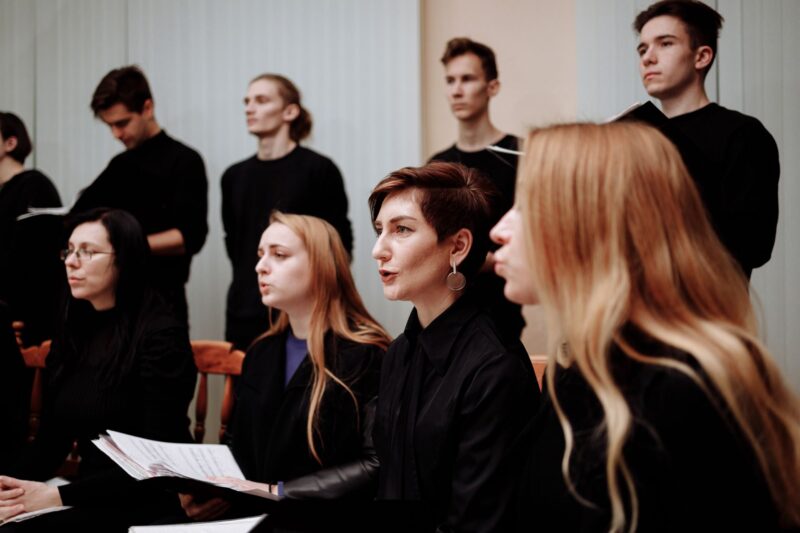 Eine Gruppe von etwa acht Personen in schwarzer Kleidung bei einer Chorprobe oder Aufführung. Im Vordergrund sitzen vier Frauen, von denen einige Notenblätter halten und singen. Dahinter stehen vier Männer, die ebenfalls in schwarzen Oberteilen gekleidet sind. Die Atmosphäre wirkt konzentriert und professionell, der Hintergrund ist hell und schlicht.