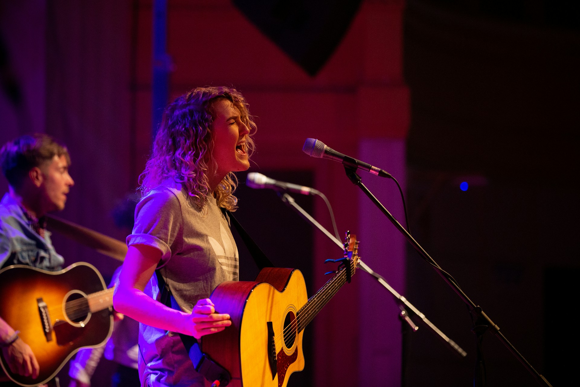 Eine Musikerin mit lockigen Haaren singt mit geschlossenen Augen leidenschaftlich in ein Mikrofon und spielt dabei eine Akustikgitarre. Sie trägt ein graues T-Shirt. Die Bühne ist in warmes violett-rotes Licht getaucht. Im Hintergrund ist ein weiterer Musiker mit einer Akustikgitarre unscharf zu erkennen.