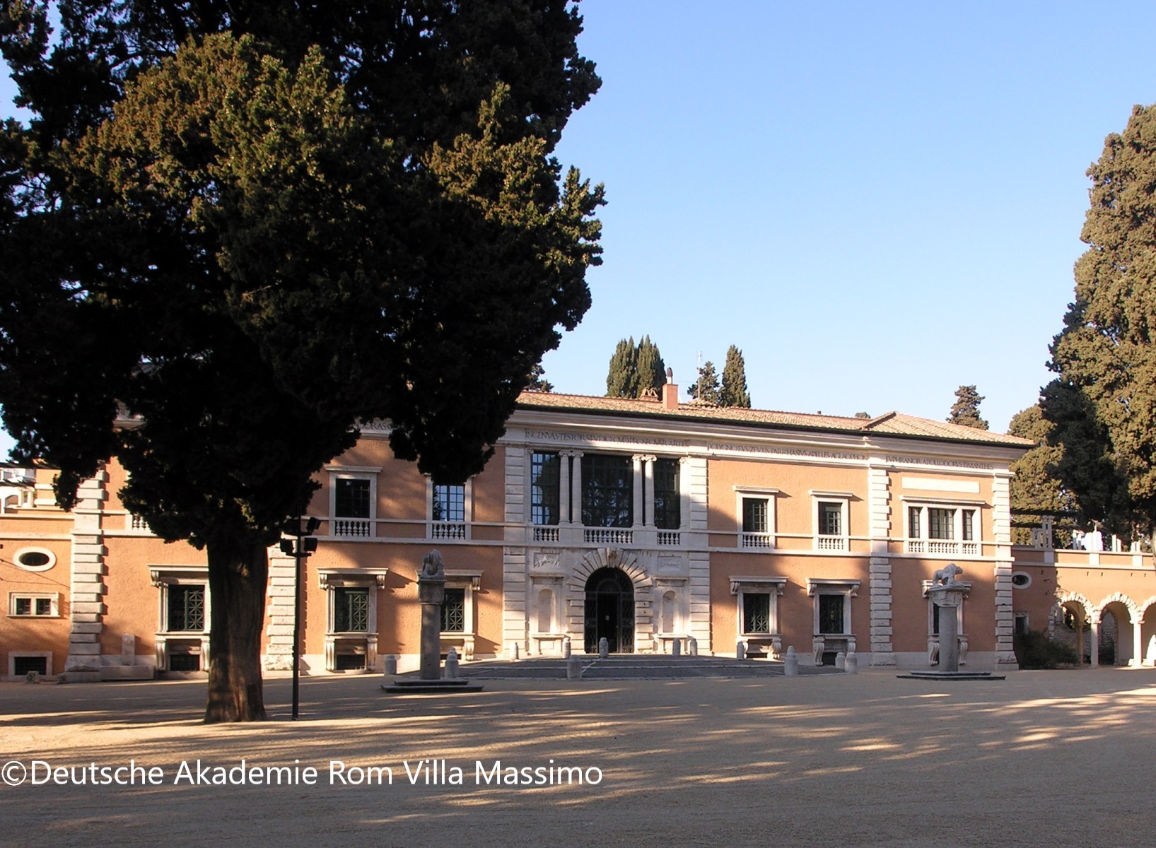 Außenansicht der Villa Massimo in Rom, dem Sitz der Deutschen Akademie Rom, bei strahlendem Sonnenschein. Das klassizistische, zweistöckige Gebäude in Terrakottarot hat eine repräsentative Fassade mit Säulenportikus, Balkonen und einem zentralen Eingangsportal. Davor liegt ein weitläufiger, gepflasterter Vorplatz mit alten Bäumen. Foto: Deutsche Akademie Rom Villa Massimo.