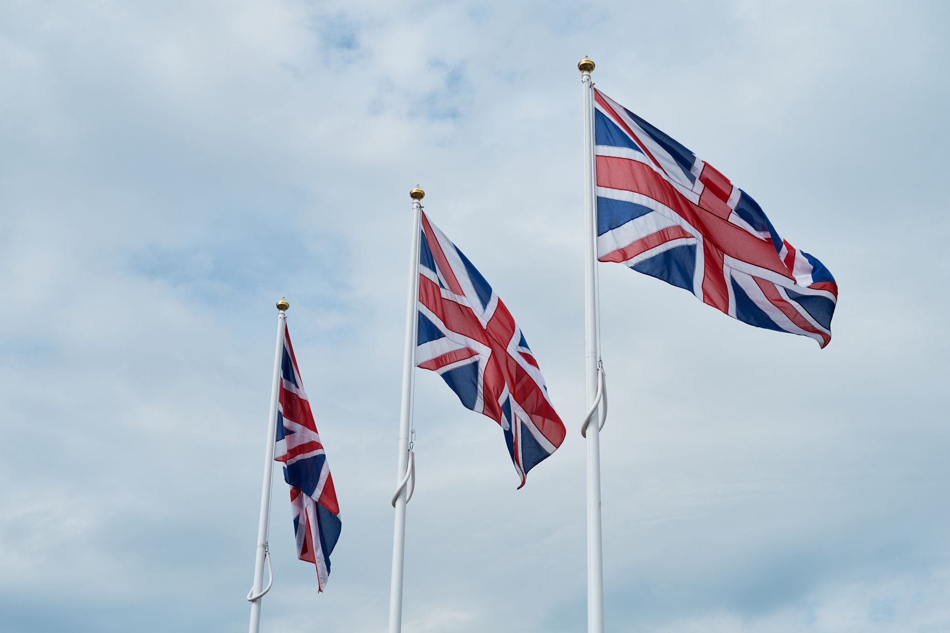 Drei britische Flaggen (Union Jack) wehen an weißen Fahnenmasten mit goldenen Knäufen vor bewölktem, hellblauem Himmel. Die Flaggen in Rot, Weiß und Blau sind durch den Wind in Bewegung, wobei die rechte Fahne am stärksten weht.