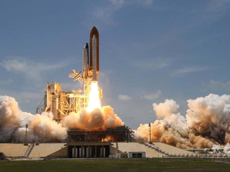 Ein Space Shuttle startet von einer Rampe in den Weltraum. Das Bild zeigt den Moment des Abhebens, mit dichten Rauch- und Feuersäulen, die aus den Triebwerken strömen. Im Hintergrund ist ein klarer blauer Himmel zu sehen.