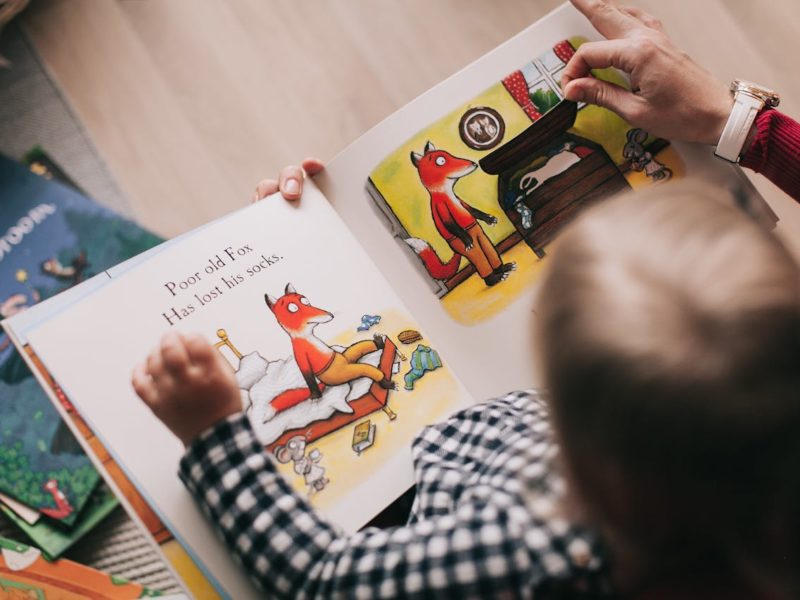 Eine Person hält ein Bilderbuch in den Händen, während ein kleines Kind auf ihrem Schoß sitzt und die Seiten betrachtet. Die Buchseite zeigt eine Illustration eines Fuchses in einem Schlafzimmer mit der Textzeile: „Poor old Fox. Has lost his socks.“ Im Hintergrund liegen weitere Kinderbücher verstreut. Die Szene vermittelt eine gemütliche Atmosphäre beim gemeinsamen Lesen.