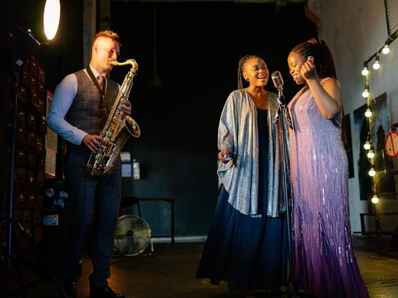 Drei Musiker:innen stehen auf einer Bühne. Links spielt ein Mann im Anzug Saxophon, während zwei Frauen in glitzernden, eleganten Kleidern rechts neben ihm singen. Die Bühne ist stimmungsvoll beleuchtet, und im Hintergrund sind Lichterketten zu sehen, die für eine warme Atmosphäre sorgen.