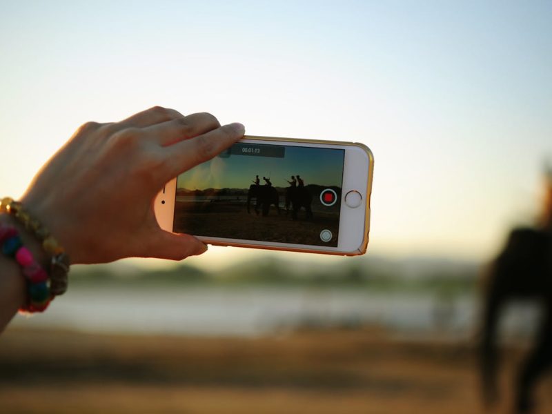 Ein Smartphone wird von einer Person mit bunten Armbändern gehalten, während ein Video aufgenommen wird. Im Display des Smartphones sind Silhouetten von Menschen zu sehen, die auf Elefanten reiten, und im Hintergrund sind Berge und ein Sonnenuntergang sichtbar.