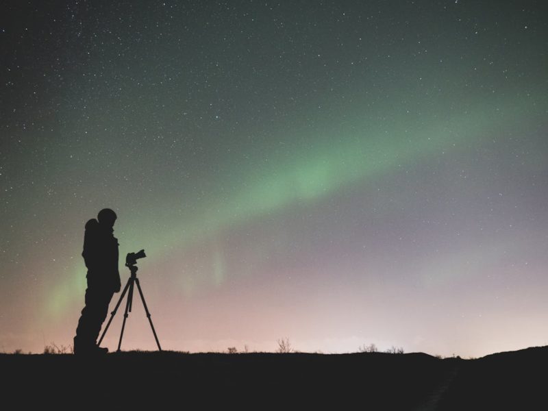 Die Grafik zeigt eine Person, die in einer dunklen, offenen Landschaft vor einem Stativ mit Kamera steht und in den Nachthimmel fotografiert. Der Himmel ist mit zahlreichen Sternen bedeckt, und es sind grünliche Lichter zu sehen, die möglicherweise das Nordlicht darstellen. Die Szene ist ruhig und vermittelt eine Atmosphäre der Stille und Naturverbundenheit, während die Person sich auf das Fotografieren des Himmels konzentriert.