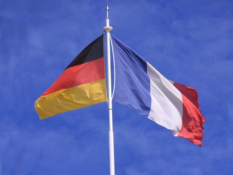 Eine deutsche und eine französische Flagge wehen nebeneinander an einem Mast vor klarem, blauem Himmel. Die deutsche Flagge zeigt die Farben Schwarz, Rot und Gold, die französische Flagge ist in Blau, Weiß und Rot gehalten.