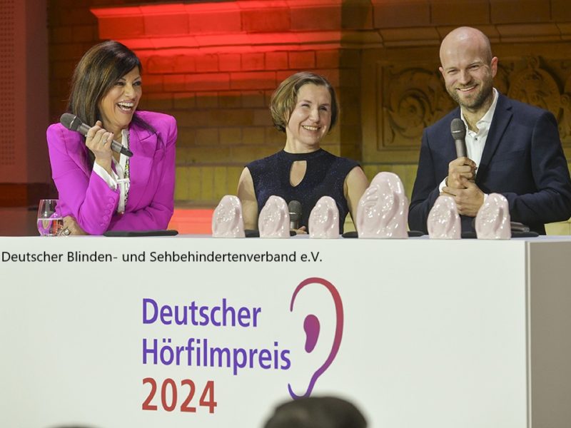 Drei Personen stehen hinter einem Podium bei der Verleihung des Deutschen Hörfilmpreises 2024. Eine Frau im pinken Blazer und zwei weitere Personen, eine Frau und ein Mann, halten Mikrofone und lächeln. Auf dem Podium steht der Schriftzug "Deutscher Hörfilmpreis 2024", daneben das Logo mit einem stilisierten Ohr. Mehrere gläserne Trophäen in Form von Ohren sind auf dem Podium ausgestellt.