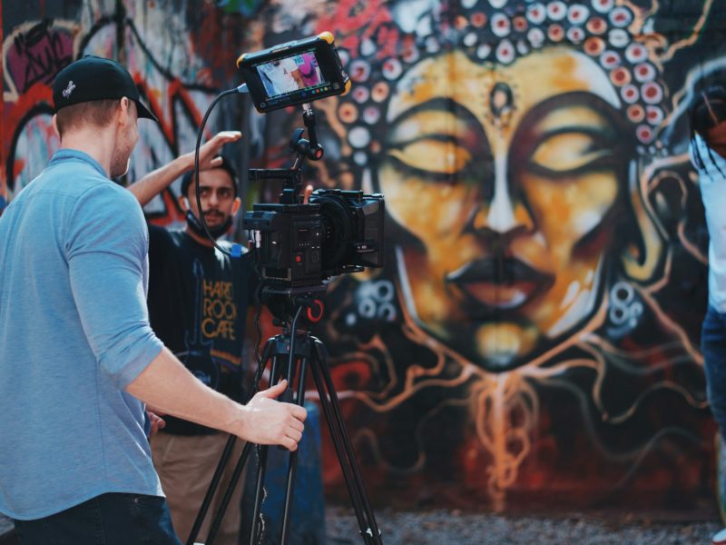 Ein Team dreht ein Video vor einer Wand mit farbenfroher Graffiti-Kunst. Im Vordergrund steht ein Kameramann mit Baseballkappe, der eine professionelle Filmkamera auf einem Stativ bedient. Im Hintergrund gibt ein weiterer Teammitglied Anweisungen und ein Model posiert neben der kunstvoll gestalteten Wand, auf der ein meditatives Gesicht mit geschlossenen Augen dargestellt ist.