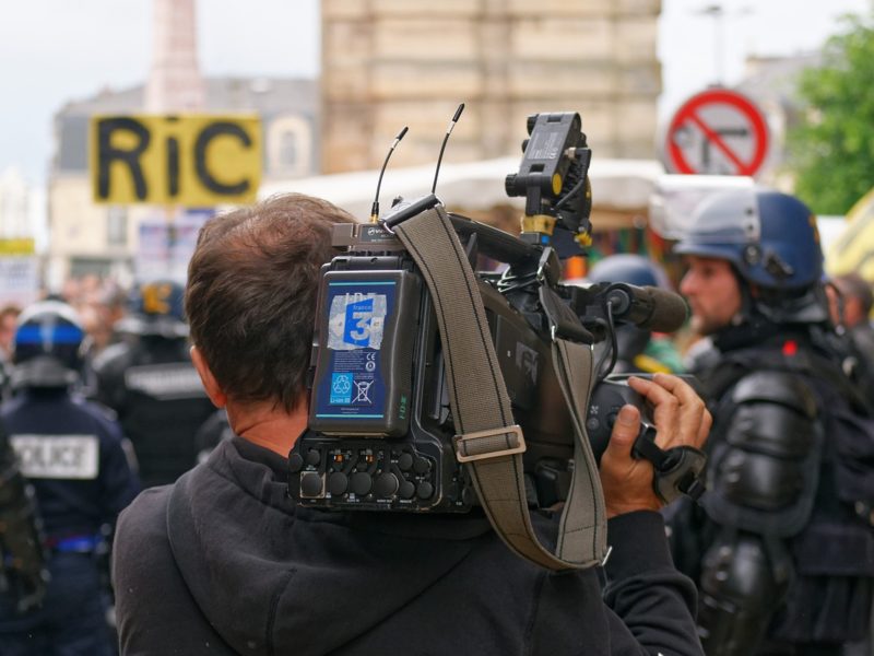 Ein Kameramann nimmt an einer Protestveranstaltung teil und filmt mit einer Schulterkamera das Geschehen. Im Vordergrund sieht man den Kameramann von hinten, während er das Geschehen aufnimmt, das sich vor ihm abspielt. Die Umgebung ist geprägt von einer Menschenmenge und mehreren Polizist in Schutzkleidung, die im Hintergrund sichtbar sind. Ein Plakat mit der Aufschrift »RIC« hebt sich ebenfalls von der Szene ab. Die Szene suggeriert eine Dokumentation von aktuellen politischen Ereignissen, vermutlich im Kontext einer Demonstration.