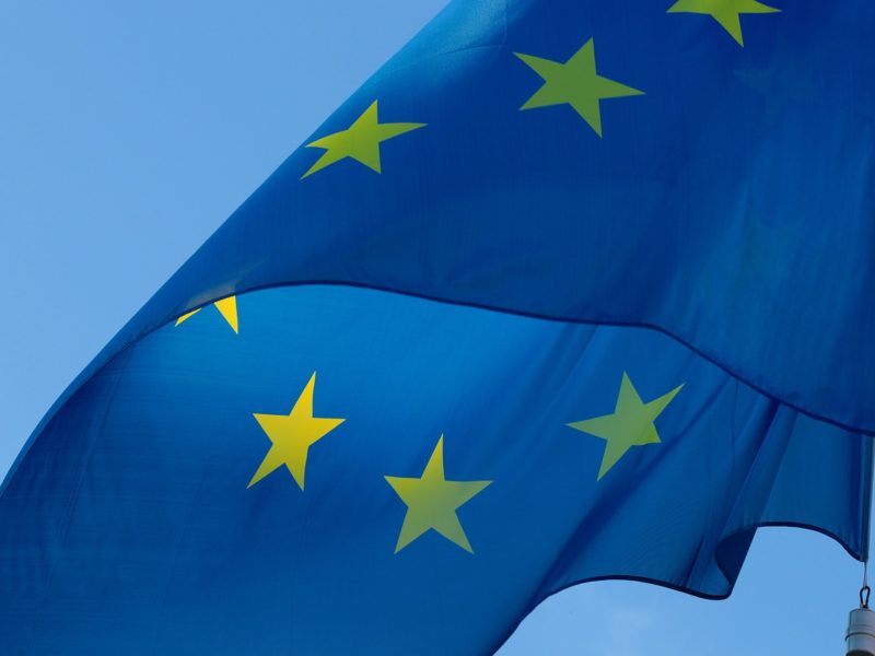 Eine Europaflagge weht im Wind vor einem blauen Himmel. Die Flagge zeigt einen blauen Hintergrund mit gelben Sternen, die im Kreis angeordnet sind und die Einheit der Europäischen Union symbolisieren.