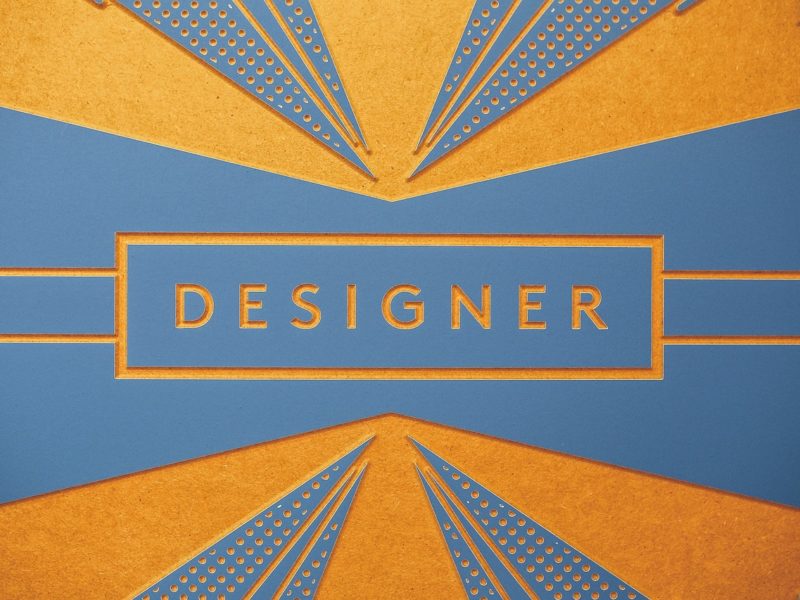 Das Bild zeigt eine stilisierte Grafik mit dem Wort "DESIGNER" in großen Buchstaben. Die Farben Blau und Orange dominieren das Design, das geometrische Muster und feine Details im Art-Deco-Stil verwendet.