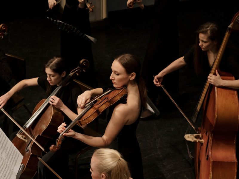 Die Grafik zeigt eine Gruppe von Musikerinnen, die Streichinstrumente spielen. Im Vordergrund sieht man eine Frau mit einer Viola, während um sie herum weitere Musikerinnen auf Cello und Kontrabass konzentriert spielen. Alle tragen formelle, schwarze Kleidung, und die Szene wirkt wie ein klassisches Konzert, das in einem geschlossenen Raum stattfindet.