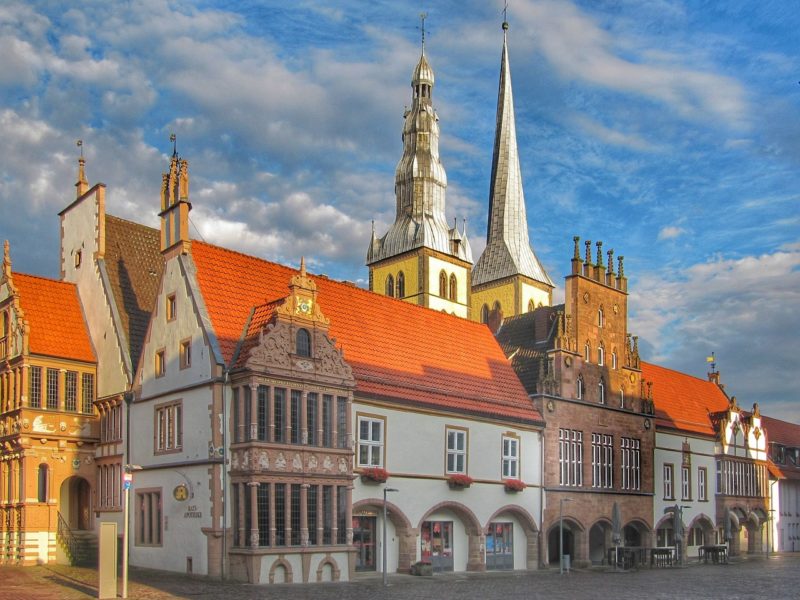 Historische Gebäude am Marktplatz in Lemgo, Deutschland, mit charakteristischen roten Ziegeldächern und verzierten Fassaden. Im Hintergrund sind die Türme einer Kirche sichtbar, die sich gegen einen blau bewölkten Himmel abheben.