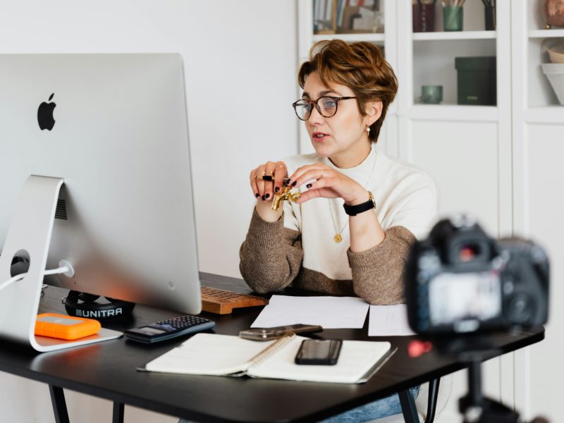 Frau mit Brille und kurzen braunen Haaren sitzt an einem Schreibtisch vor einem großen Computerbildschirm. Sie trägt einen Pullover in Creme und Braun und hält einen goldenen Stift in den Händen. Auf dem Schreibtisch liegen ein Notizbuch, ein Smartphone, eine externe Festplatte, ein Taschenrechner und einige Blätter Papier. Im Vordergrund steht eine Kamera auf einem Stativ, die auf sie gerichtet ist. Im Hintergrund sind Regale mit Büchern und Dekoration zu sehen.