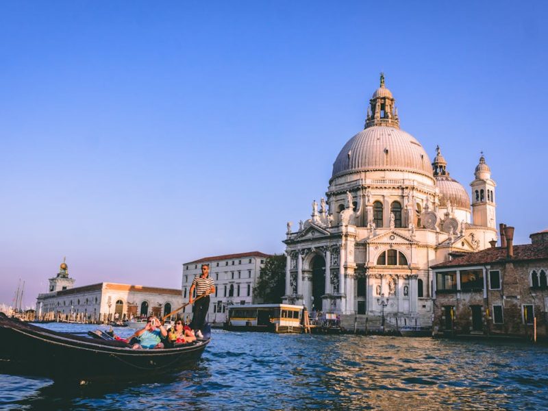 Eine klassische venezianische Gondel mit mehreren Passagier und einem Gondoliere fährt auf dem Wasser vor der imposanten Basilika Santa Maria della Salute in Venedig. Die Abendsonne beleuchtet die historische Architektur und die Wasseroberfläche, während ein wolkenloser, blauer Himmel die Szene umrahmt.