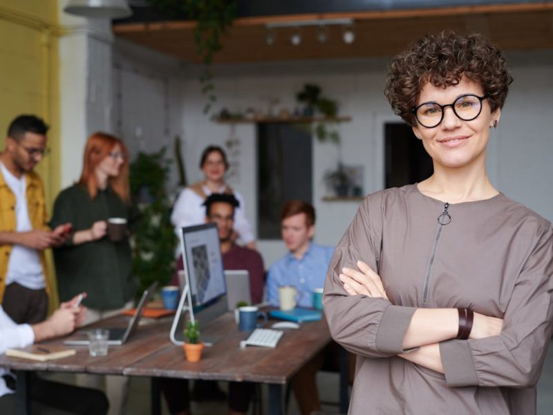 Eine lächelnde Frau mit lockigem Haar und Brille steht im Vordergrund eines modernen Büro-Settings mit verschränkten Armen. Im Hintergrund arbeitet eine Gruppe von Menschen an einem Tisch mit Laptops, Pflanzen und Notizbüchern, während sie miteinander interagieren.