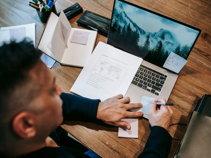 Eine Person arbeitet konzentriert an einem Schreibtisch mit einem Laptop, mehreren Notizbüchern und Dokumenten. Auf dem Laptopbildschirm ist eine Naturaufnahme mit Bergen zu sehen. Die Person macht sich handschriftliche Notizen auf einem Zettel und hat weitere Notizzettel auf dem Laptop befestigt. Der Arbeitsplatz wirkt gut organisiert, mit Schreibutensilien und Papieren.