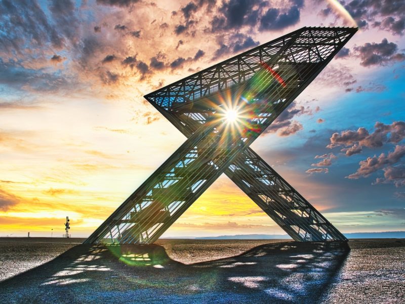 Ein modernes Stahlmonument in X-Form, das Saar-Polygon, steht vor einem beeindruckenden Sonnenuntergang, wobei Sonnenstrahlen durch die Struktur brechen. Im Hintergrund sind Wolken am farbenfrohen Himmel und eine weite, offene Landschaft zu sehen.