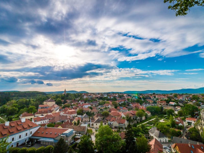 Panorama einer kleinen Stadt mit roten Ziegeldächern, umgeben von grünen Wäldern und Hügeln unter einem blauen Himmel mit weißen Wolken. Im Hintergrund sind eine Kirche mit Turm und eine weite, malerische Landschaft zu sehen.