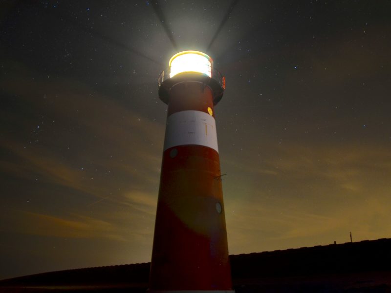 Ein rot-weiß gestreifter Leuchtturm leuchtet in die Dunkelheit und strahlt Lichtstrahlen in den Nachthimmel. Sterne sind im Hintergrund sichtbar, und der Himmel ist leicht bewölkt, was eine mystische Atmosphäre erzeugt.