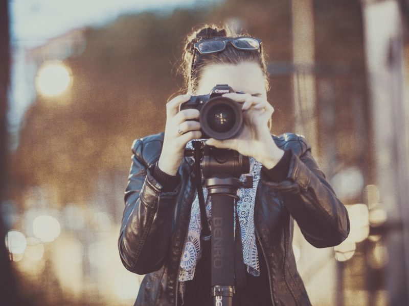 Eine Fotografin steht hinter ihrer Kamera und richtet den Fokus auf ein Motiv. Sie trägt eine Lederjacke und hält die Kamera mit beiden Händen. Im Hintergrund sind unscharfe Lichter und eine städtische Umgebung bei Dämmerung zu sehen. Die Szene vermittelt Kreativität und Leidenschaft für die Fotografie.