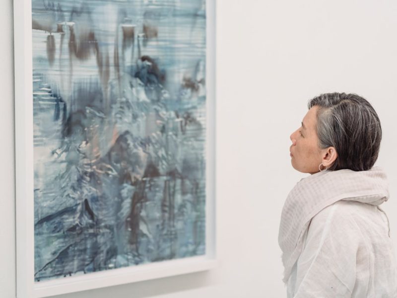 Eine Frau mit grauem, kurzem Haar betrachtet ein abstraktes Gemälde in blauen und grauen Farbtönen in einer Kunstgalerie. Sie trägt einen hellen Schal und eine weiße, lockere Bluse. Das Bild hat eine ruhige und nachdenkliche Atmosphäre.