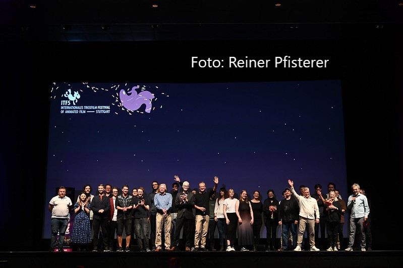 Eine Gruppe von Menschen steht auf der Bühne des Internationalen Trickfilm-Festivals Stuttgart (ITFS) vor einem großen Bildschirm mit dem Festival-Logo. Die Personen, darunter mehrere Filmschaffende und Veranstalter:innen, lächeln, klatschen oder winken ins Publikum. Oben rechts im Bild ist der Hinweis »Foto: Reiner Pfisterer« zu sehen.