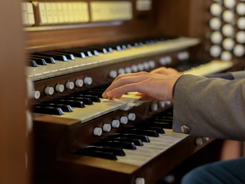 Eine Nahaufnahme von Händen, die auf den Tasten einer Kirchenorgel spielen. Die Orgel hat mehrere Tastenreihen und zahlreiche Knöpfe zur Klangsteuerung.