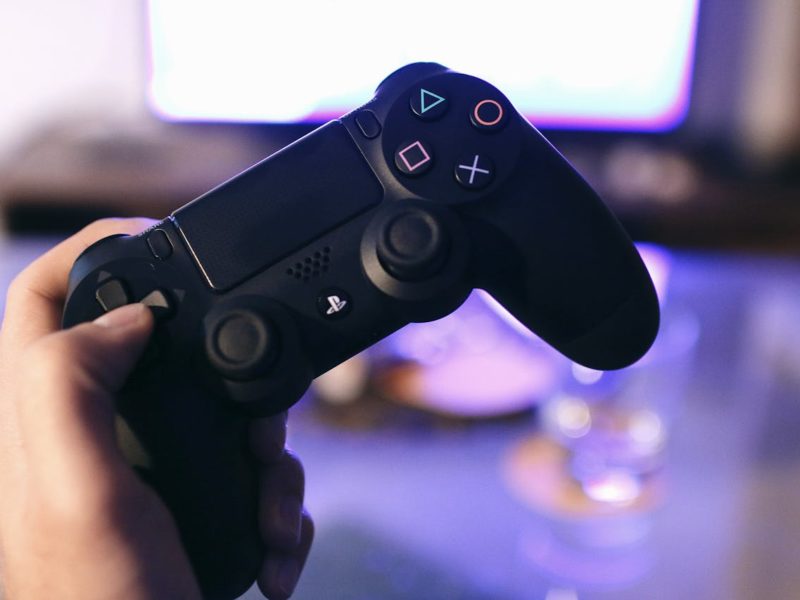 Eine Hand hält einen schwarzen Gaming-Controller, der deutlich die farbigen Tasten und Analogsticks zeigt. Im Hintergrund ist ein unscharfer Bildschirm mit violettem Licht zu erkennen, was eine entspannte Gaming-Atmosphäre andeutet.