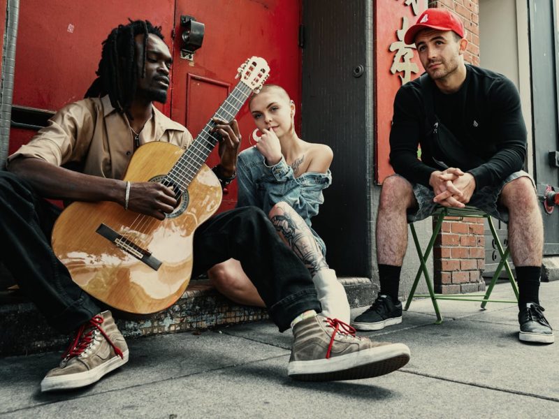 Drei junge Menschen sitzen vor einer roten Tür auf einer Straße. Der Mann links, mit dunklen Dreadlocks und einem beigen Hemd, spielt eine akustische Gitarre. Die Frau in der Mitte, mit rasiertem Kopf, großen Ohrringen und einem Jeans-Oberteil, sitzt lässig auf dem Boden und schaut nachdenklich. Der Mann rechts, in einem schwarzen Pullover, Camouflage-Shorts und einer roten Baseballkappe, sitzt auf einem grünen Klappstuhl und beobachtet die Szene entspannt. Die Atmosphäre wirkt urban und kreativ.