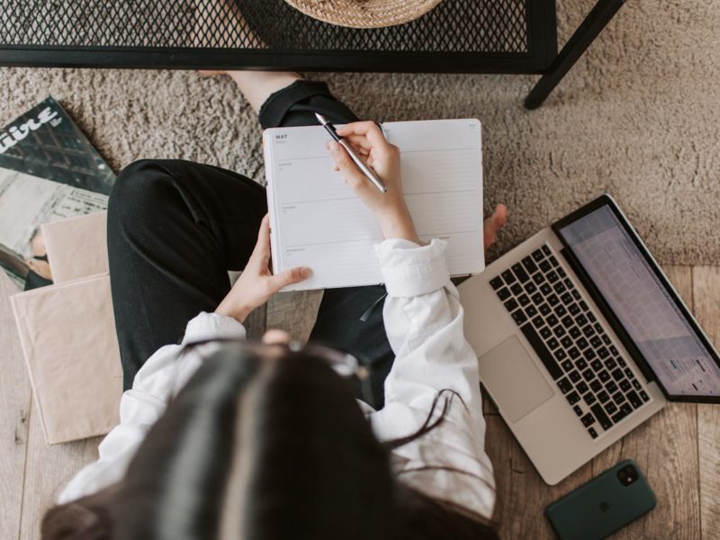 Eine Frau sitzt auf dem Boden neben einem Couchtisch und arbeitet. Sie hält einen Stift in der Hand und schreibt in einen offenen Planer. Neben ihr liegt ein aufgeklappter Laptop, ein Smartphone und einige Bücher. Der Raum hat einen Holzboden mit einem Teppich, und eine Zeitschrift ist ebenfalls sichtbar.