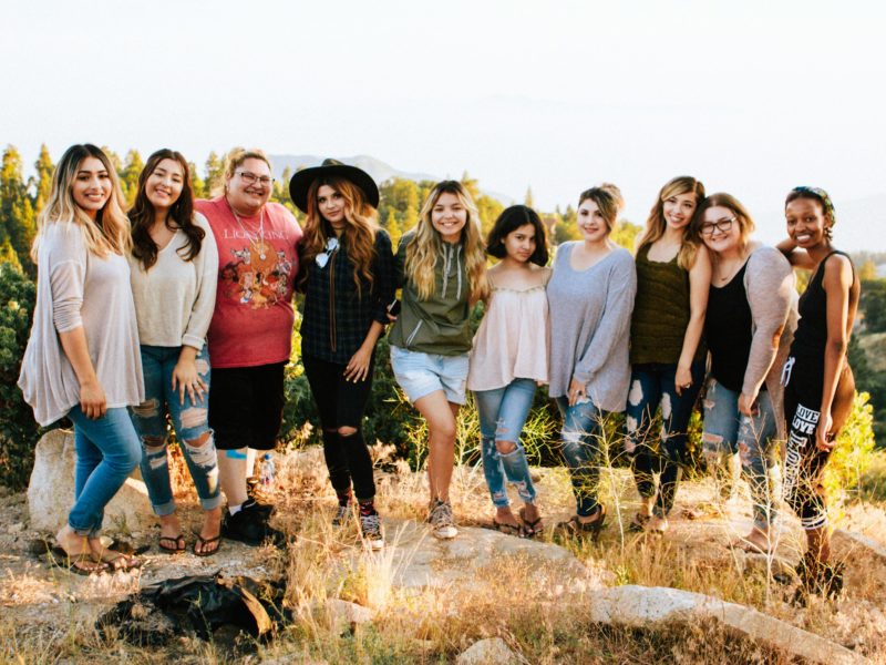 Eine Gruppe von zehn Frauen steht auf einem sonnigen Hügel inmitten einer natürlichen Landschaft. Sie lächeln fröhlich in die Kamera, umgeben von Büschen und Bäumen im Hintergrund.