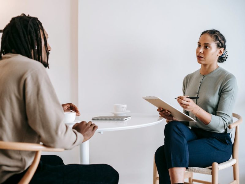 Eine Frau mit dunklen, geflochtenen Haaren sitzt an einem runden Tisch und führt ein Gespräch mit einem Mann mit Dreadlocks. Sie hält ein Klemmbrett und einen Stift in der Hand, während er eine Tasse Kaffee vor sich hat. Beide wirken konzentriert. Im Hintergrund ist eine weiße Wand mit einem gerahmten Bild zu sehen.