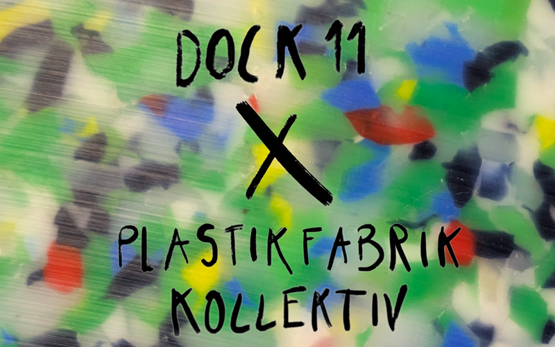 Nahaufnahme einer recycelten Plastikplatte mit bunten Farbpartikeln in Grün, Blau, Rot, Gelb und Schwarz. Darauf steht in handgeschriebener Schrift: »DOCK 11 X PLASTIKFABRIK KOLLEKTIV«.
