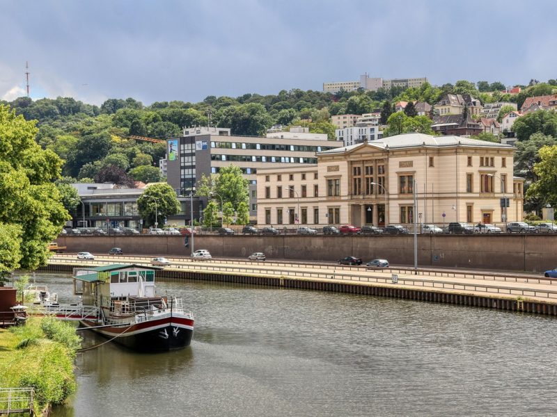 Blick auf das Saarufer in Saarbrücken mit dem saarländischen Landtag im Vordergrund, einer Uferstraße mit Autos und einem angelegten Boot auf der Saar. Im Hintergrund Wohnhäuser und bewaldete Hügel.