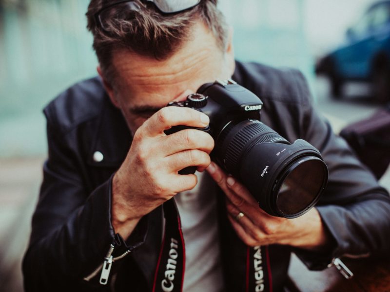 Ein Mann in Lederjacke hält eine Canon-Spiegelreflexkamera mit Sigma-Objektiv vor sein Gesicht und macht ein Foto; er sitzt im Freien und trägt eine Sonnenbrille auf dem Kopf.