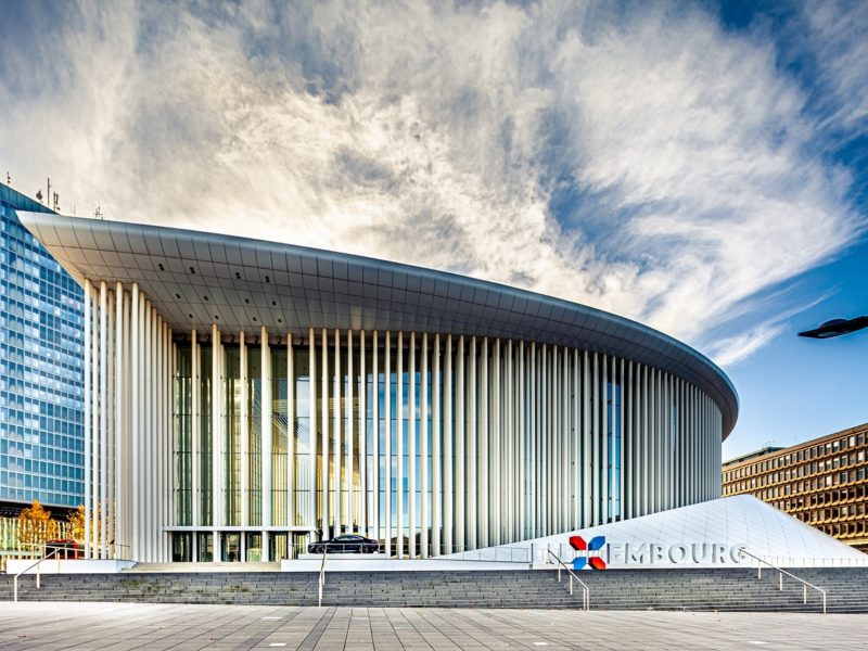 Außenansicht der Philharmonie Luxemburg mit ihrer modernen Architektur, großen Glasfassade und markanten weißen Säulen vor blauem Himmel.
