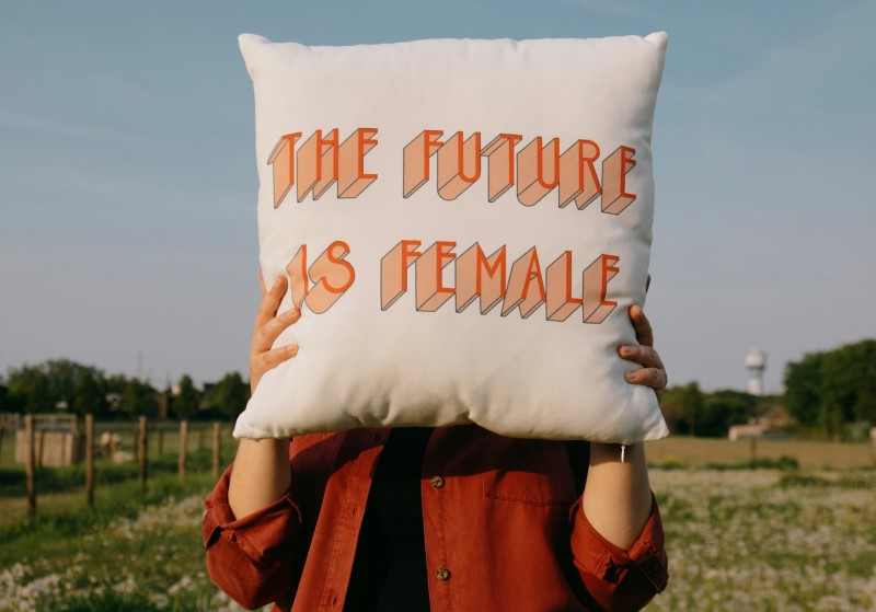 Eine Person steht auf einer Wiese und hält ein weißes Kissen vor ihr Gesicht. Auf dem Kissen steht in orange-roten Buchstaben: "THE FUTURE IS FEMALE".