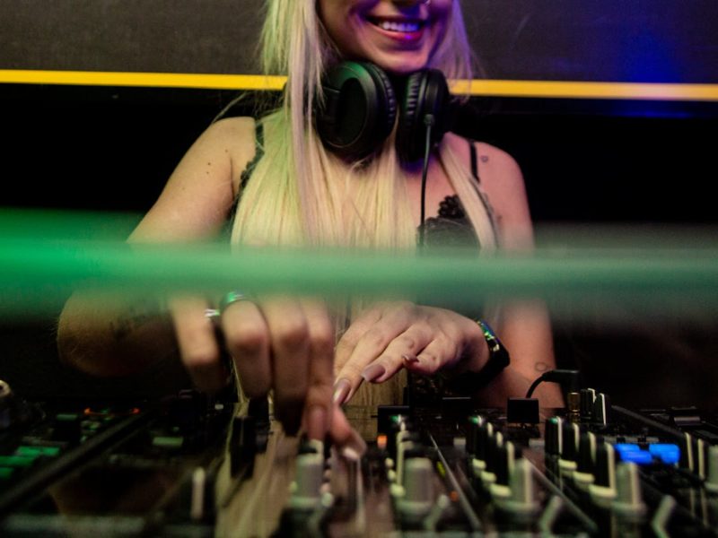Eine junge DJane mit langen blonden Haaren und Kopfhörern um den Hals steht an einem Mischpult und legt auf. Sie lächelt, während sie konzentriert an den Reglern arbeitet. Das Foto zeigt eine Nahaufnahme ihrer Hände und des Mischpults in einem clubähnlichen Setting mit farbigen Lichtern.