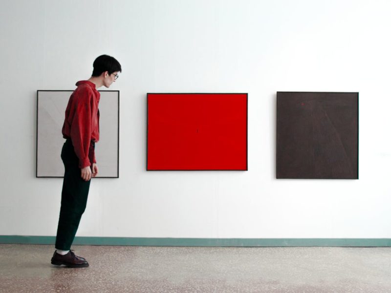 Eine Person in rotem Hemd und schwarzer Hose betrachtet in einer Galerie drei abstrakte, quadratische Gemälde an einer weißen Wand – eines weiß, eines rot und eines dunkelbraun.