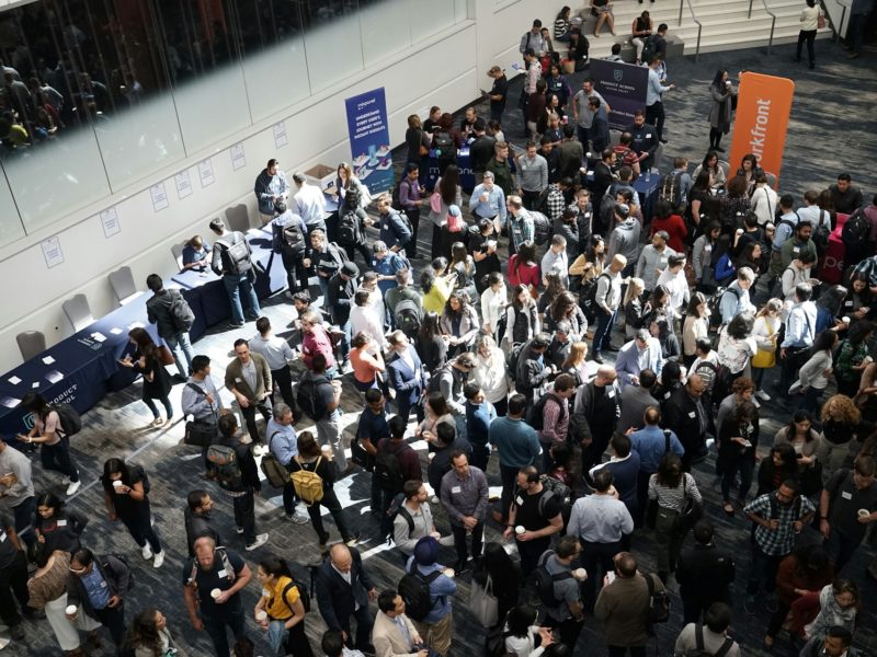 Großveranstaltung in einem modernen Konferenzzentrum mit zahlreichen Menschen, die sich an Infoständen und in Gruppen unterhalten. Die Szene zeigt ein reges Networking-Event mit Banner-Displays und Namensschildern.