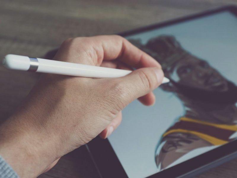 Eine Hand hält einen digitalen Eingabestift und zeichnet auf einem Tablet ein Porträt eines Mannes mit Bart und Basketballtrikot.