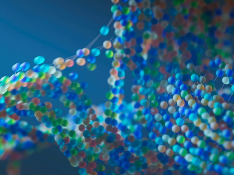 Bunte, unscharf dargestellte Molekülstruktur auf blauem Hintergrund; zahlreiche kleine, farbige Kugeln sind durch dünne Verbindungen zu einem komplexen, netzartigen Muster verbunden, das an eine DNA-Doppelhelix erinnert.