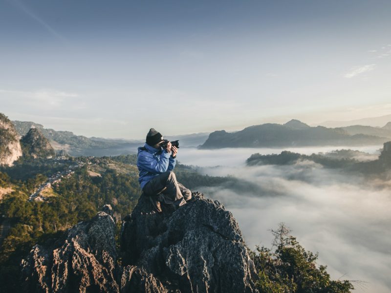 Eine Person in Outdoor-Kleidung sitzt auf einem felsigen Berggipfel und fotografiert mit einer Kamera. Im Hintergrund erstreckt sich eine nebelverhangene Hügellandschaft mit bewaldeten Bergen unter einem klaren Himmel.